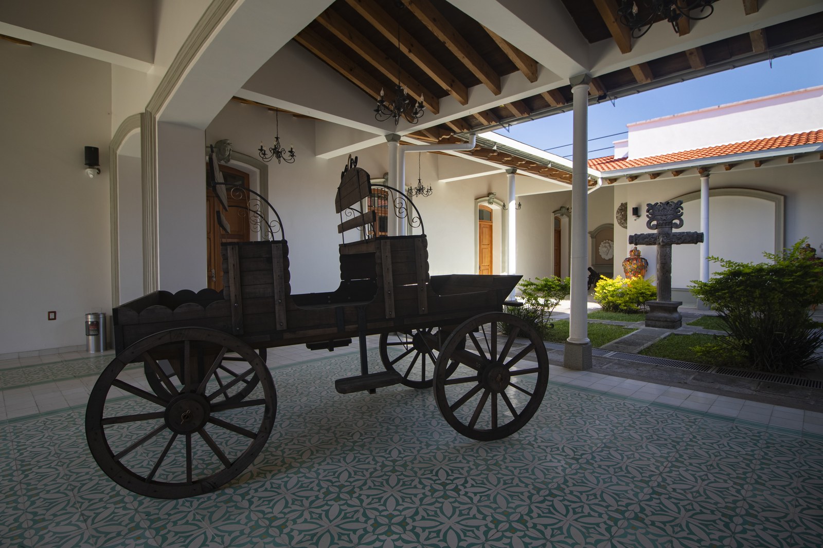 Wooden cart under the corridor of Casa de los Leones, on patterned tile flooring, with the courtyard and stone cross behind